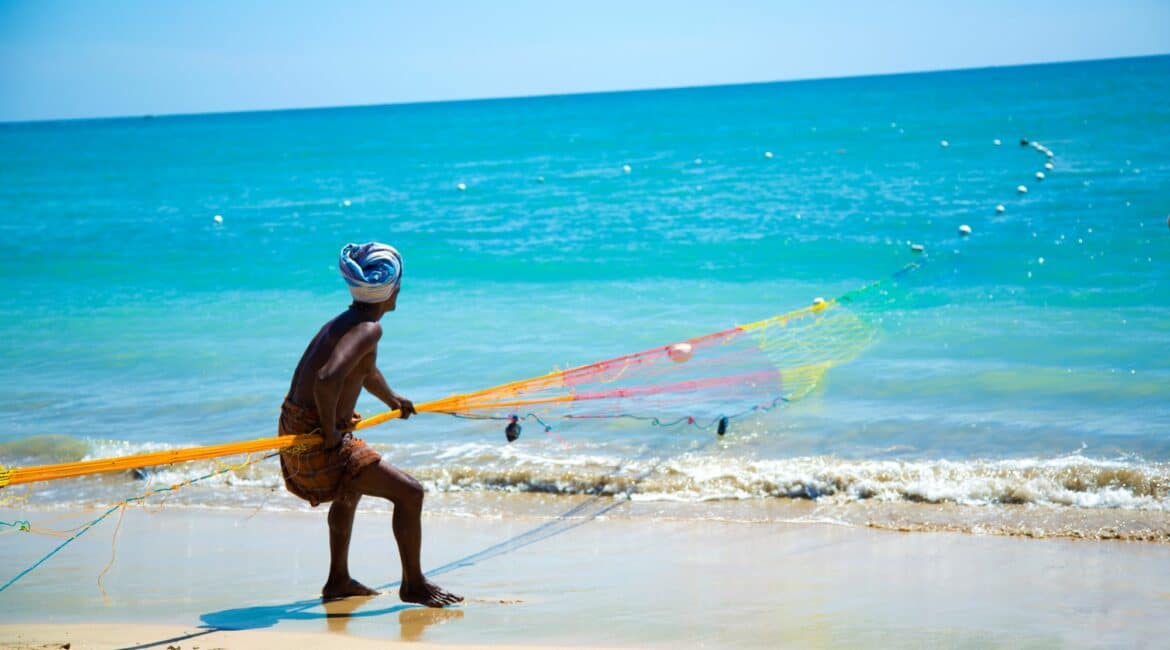 Sri Lankan fisherman hauling nets on a white sand beach beneath a bright blue sky, with the azure Indian Ocean stretching out behind him.
