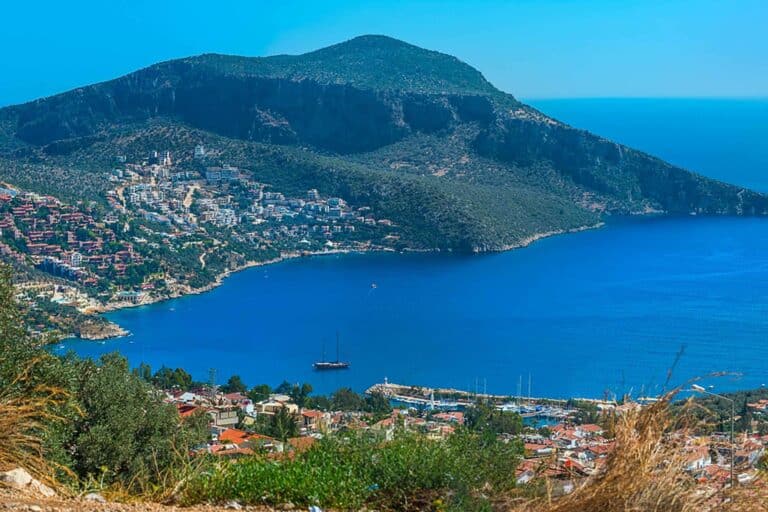 Aerial view of Kalkan Bay with gulets bobbing in the dark indigo sea, framed by a horseshoe-shaped coastline dotted with whitewashed villas and boutique hotels.