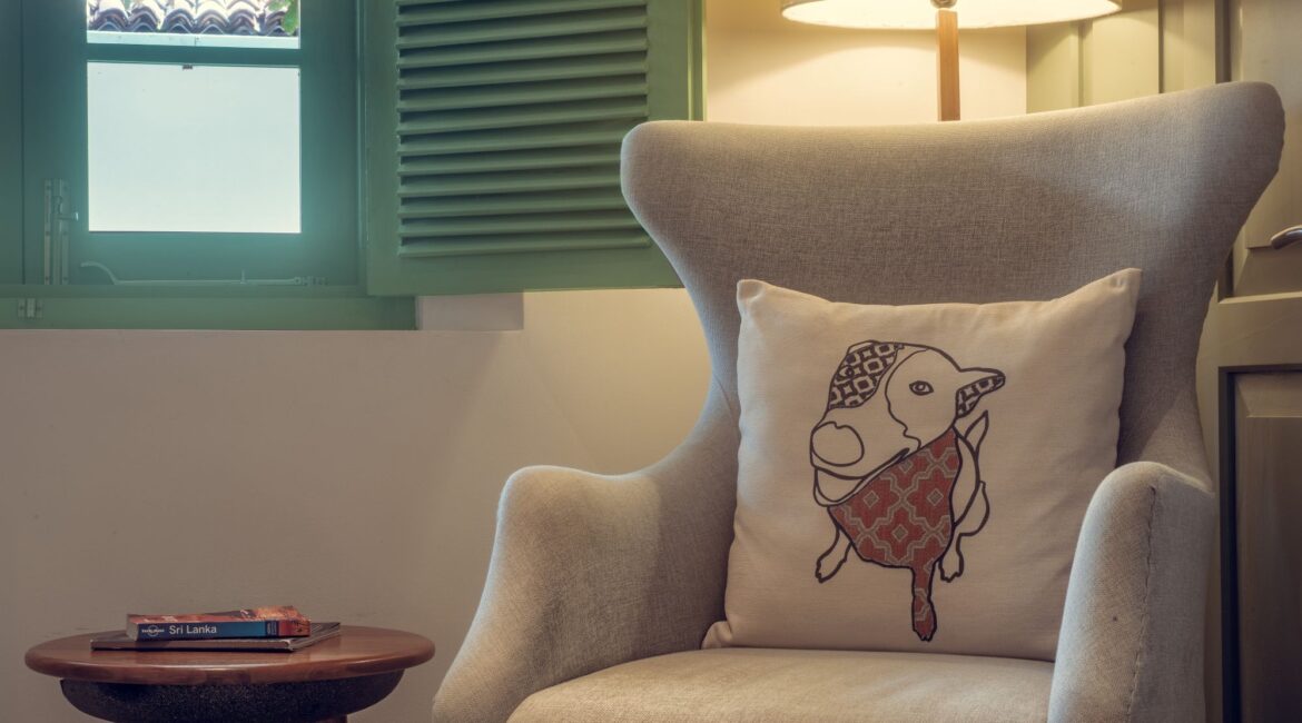 Bazaar Bedroom at Fort Bazaar Hotel Sri Lanka featuring a cosy upholstered armchair beside a wooden side table and large shuttered window.