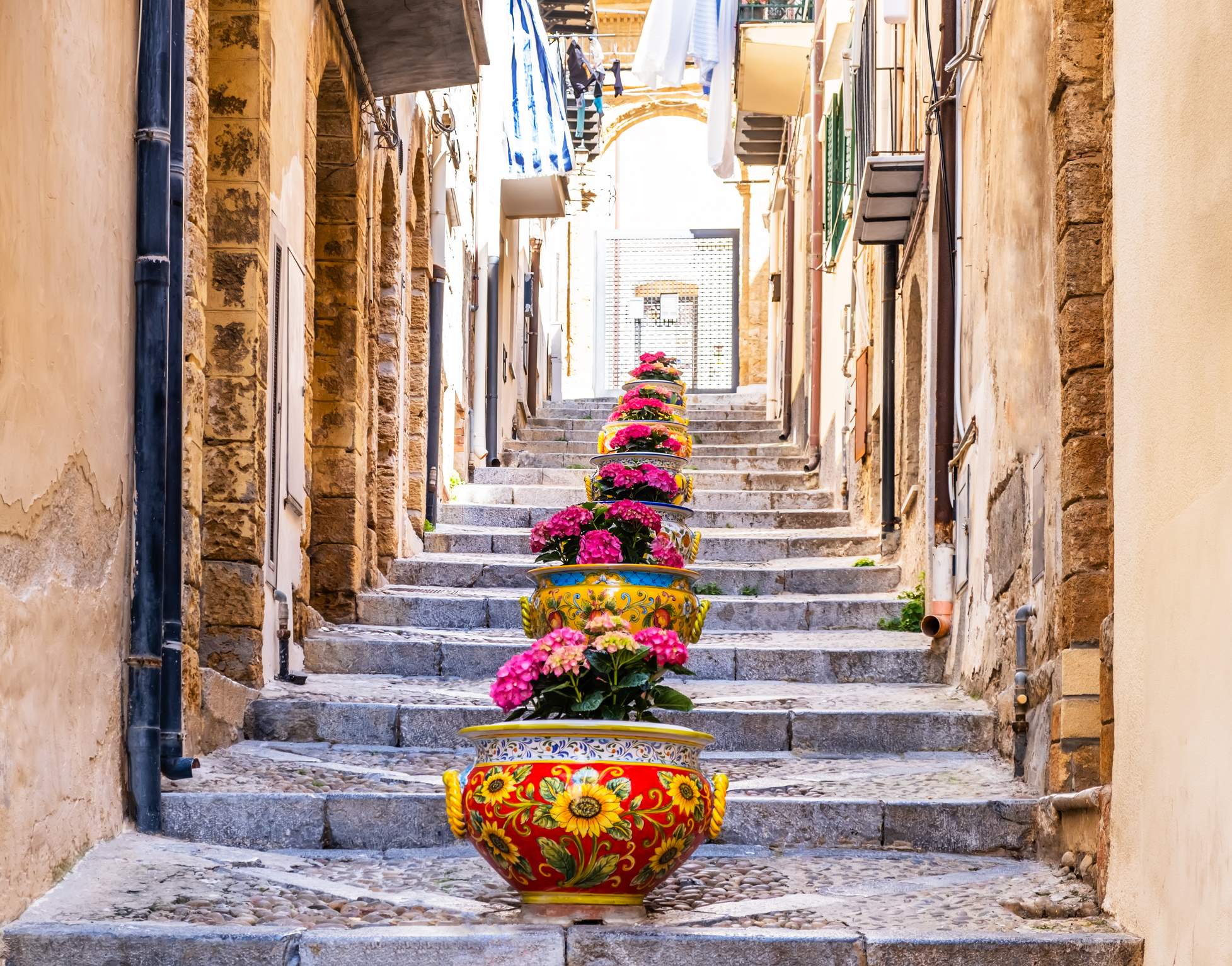 Narrow street in the old town of Cefalu, medieval village on Sicily island, Italy. Flower pots with traditional sicilian decoration. La Rocca cliff. Popular tourist attraction in Province of Palermo.